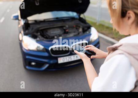 Una donna al cofano aperto di un'auto rotta chiama un rimorchio su un telefono cellulare Foto Stock