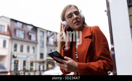 La giovane donna bionda con un telefono cellulare in mano ascolta la musica con le cuffie sullo sfondo del paesaggio urbano Foto Stock