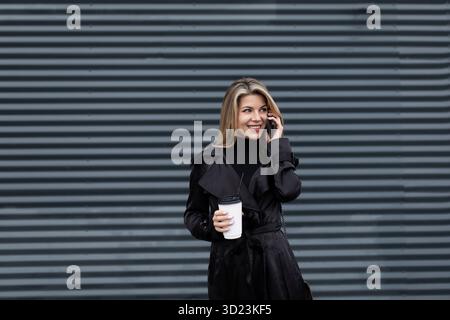 Ritratto di una dirigente donna in abiti da lavoro neri con un bicchiere di caffè che parla al telefono contro un muro scuro Foto Stock