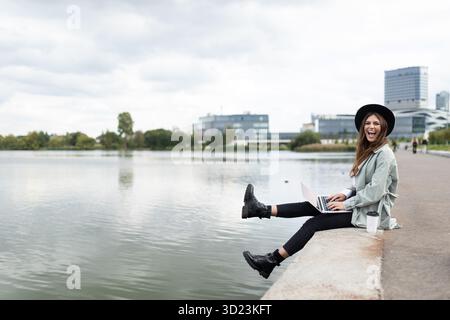 Ritratto di una giovane donna elegante e maliziosa con un ampio sorriso sulle mani sedute sul terrapieno che penzolava le gambe al Foto Stock