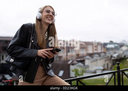 Una giovane studentessa ascolta musica piacevole sullo sfondo del paesaggio urbano con le cuffie con un telefono in mano Foto Stock