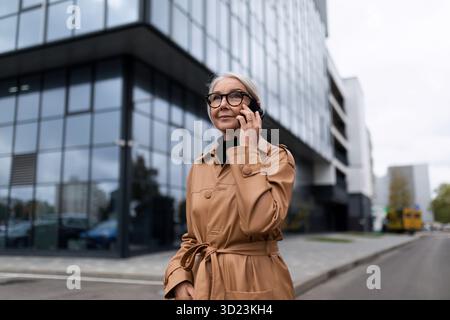 Donna caucasica di alto livello che parla al telefono sullo sfondo di un edificio di uffici Foto Stock