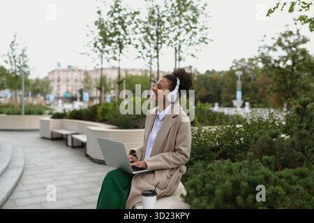Una giovane studentessa nera con un laptop seduto su una panchina che ascolta la musica con le cuffie con piacere chiudendo gli occhi Foto Stock