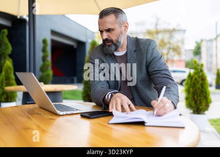 Uomo d'affari adulto che lavora online su un portatile in un bar della città vicino a un edificio di uffici, lavoro libero e lavoro remoto Foto Stock