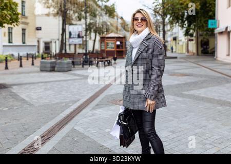 Donna elegante in abiti autunnali che cammina per la città con gli occhiali da sole Foto Stock