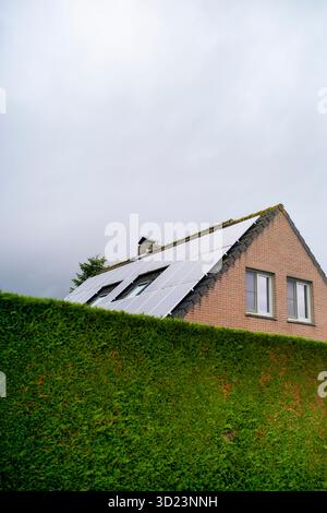 Casa in mattoni con pannelli solari sul tetto dietro una lussureggiante siepe verde sotto il cielo nuvoloso. Blankenberge, Fiandre occidentali, Belgio Foto Stock