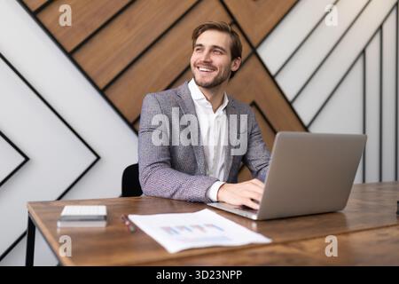 Un uomo d'affari sorridente che lavora sul laptop e guarda fuori dalla finestra Foto Stock