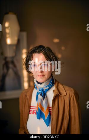 Una giovane donna che indossa occhiali e sciarpa si stende all'interno con luce soffusa e sfondo neutro. California, Stati Uniti Foto Stock