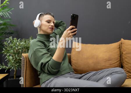 Donna matura con capelli corti sdraiata sul divano che ascolta musica con le cuffie guardando lo smartphone Foto Stock