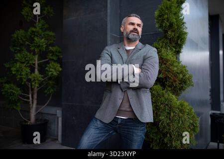 Un uomo d'affari anziano serio con una giacca grigia accanto all'ingresso dell'edificio degli uffici guarda in lontananza nel pensiero Foto Stock