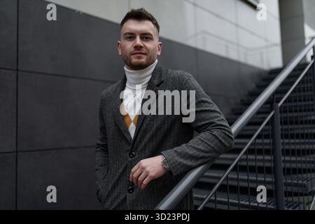 Ritratto di un uomo forte e brutale in un cappotto grigio sullo sfondo della facciata dell'edificio con una scala che porta in alto Foto Stock