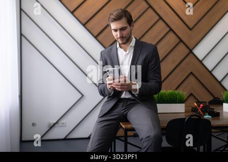 Uomo d'affari seduto sulla scrivania in ufficio a leggere messaggi al telefono Foto Stock