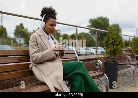 Giovane donna afroamericana che ascolta musica con le cuffie dal telefono seduto su una panchina in un parco cittadino, lavora come freelance Foto Stock