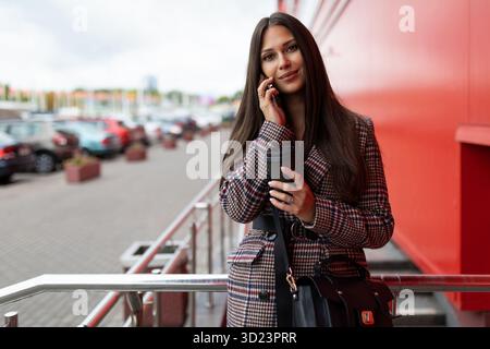 Una giovane imprenditrice di successo parla su un telefono cellulare sullo sfondo di una facciata rossa di un edificio Foto Stock