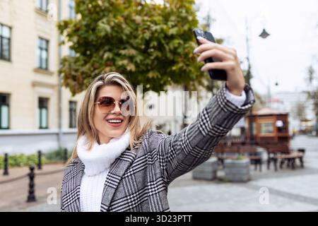 Una donna adulta con un maglione bianco e occhiali da sole fa un selfie al telefono sullo sfondo della città Foto Stock