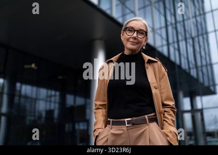 Una donna d'affari anziana sullo sfondo di un edificio di uffici guarda la telecamera con un sorriso Foto Stock
