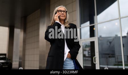 Un'anziana donna d'affari entra in ufficio parlando al telefono cellulare, donna leader nel concetto di business Foto Stock