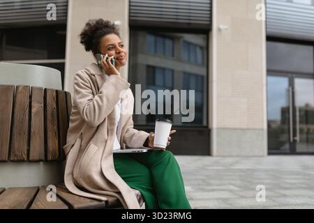 Donna d'affari afroamericana che parla al telefono cellulare e beve un caffè durante la pausa pranzo all'aperto Foto Stock