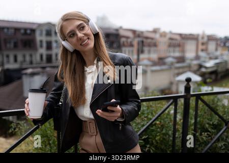 Ritratto di una giovane donna allegra e felice con un telefono cellulare e una tazza di caffè in mano che ascolta la musica attraverso la stufa Foto Stock