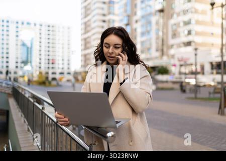 Giovane donna d'affari di successo che lavora su un notebook online che parla su un telefono cellulare in città Foto Stock