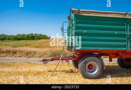 Un rimorchio agricolo verde parcheggiato in un campo di grano dorato sotto un cielo azzurro. Foto Stock