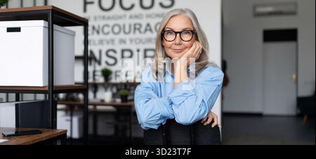 Ritratto di una donna d'affari di successo in occhiali da vista seduta su una sedia nel mezzo dell'ufficio, caucasica di mezza età Foto Stock