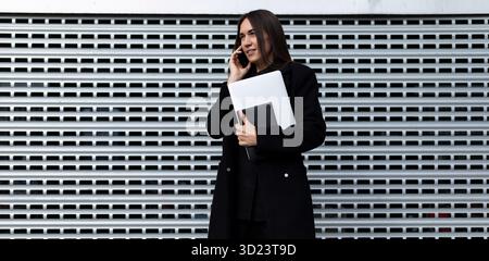 Una donna d'affari di successo con un portatile in mano parla al telefono contro una parete grigia Foto Stock
