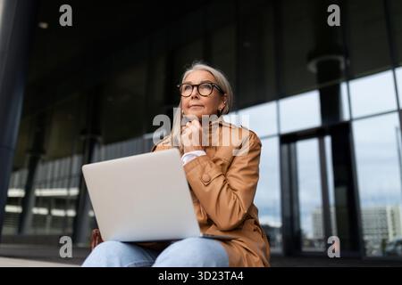 Una donna d'affari siede con un portatile in ginocchio sui gradini all'ingresso di un edificio per uffici, lavora fuori dall'ufficio Conc Foto Stock
