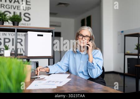 Una donna di cinquant'anni prepara i documenti seduti in ufficio a parlare al telefono cellulare. Accou. Professionale Foto Stock