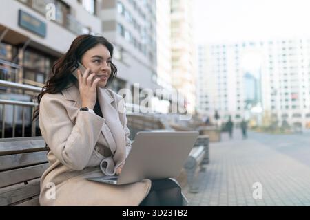 Giovane donna d'affari che parla al telefono cellulare e lavora al computer portatile online fuori dall'ufficio Foto Stock