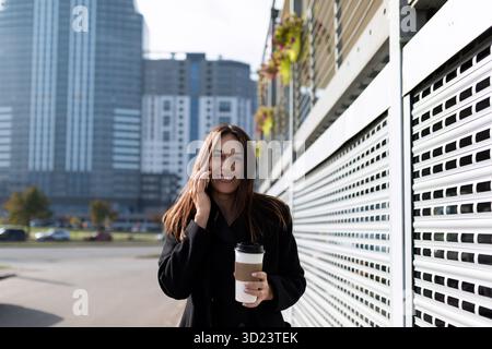 Un'elegante donna d'affari va al lavoro a parlare al telefono cellulare con una tazza di caffè Foto Stock