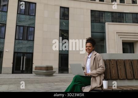 Attraente studentessa afroamericana seduta su una panchina accanto a un edificio cittadino Foto Stock