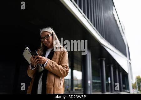 Una donna d'affari anziana di mezza età guarda il telefono con un portatile tra le mani accanto al centro ufficio, lavora fuori dal Foto Stock