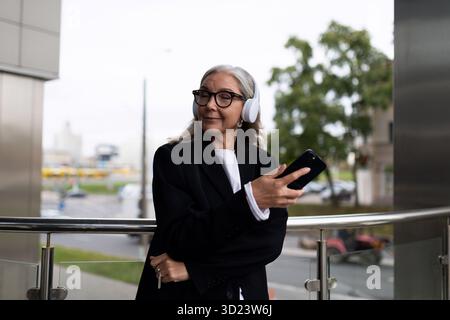 Donna anziana ed elegante che ascolta con piacere la musica con le cuffie con il telefono cellulare nelle mani fuori dall'ufficio Foto Stock