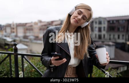 La giovane donna bionda, che guarda le mani del telefono cellulare e un bicchiere di caffè, ascolta la musica con le cuffie chiudendo gli occhi con piacere Foto Stock