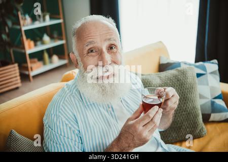 Un allegro pensionato si rilassa a casa gustando una tazza di tè su un accogliente divano in un soggiorno soleggiato, creando una calda e confortevole atmosfera domestica Foto Stock