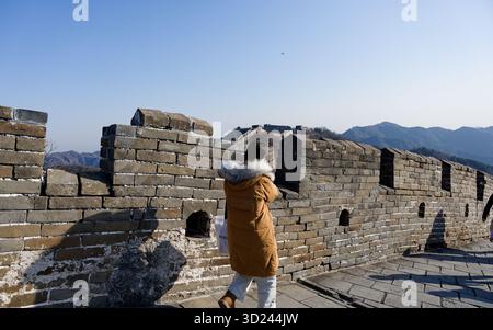 Il visitatore con un lungo cappotto invernale marrone cammina lungo l'antico sentiero della grande Muraglia, incorniciato da montagne lontane e da un pallido cielo invernale Foto Stock