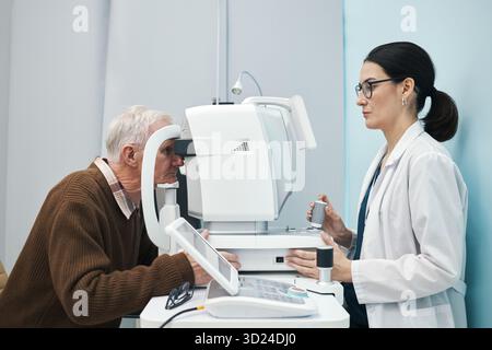 Uomo caucasico anziano sottoposto a esame oculistico con dispositivo oftalmico operato da giovane medico donna caucasica adulta, entrambi concentrati sulla procedura in ambiente clinico Foto Stock
