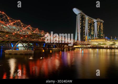 Vista dell'Helix Bridge illuminato di notte con Marina Bay Sands in lontananza che si riflette sull'acqua, Singapore, Singapore. Foto Stock