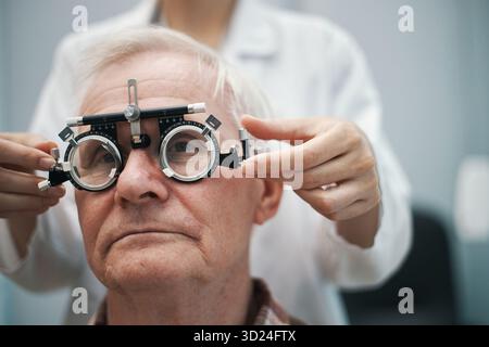 Uomo caucasico anziano sottoposto a esame oculistico con optometrista che regola il phoropter durante il test di visione in clinica medica, soggetto che guarda dritto avanti, primo piano del viso e del dispositivo Foto Stock