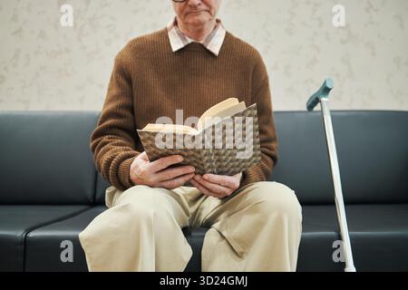 Uomo caucasico anziano seduto sul divano nella sala d'attesa leggendo un libro con copertina a motivi geometrici, tenendo le pagine aperte con entrambe le mani, bastone da passeggio che riposa accanto, indossando occhiali e abbigliamento casual Foto Stock