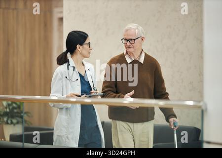 Donna asiatica di mezza età medico che cammina e parla con l'uomo caucasico anziano che tiene il bastone in struttura medica, entrambi impegnati in conversazione, medico che tiene il tablet digitale Foto Stock