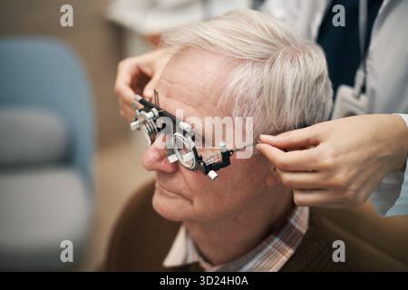 Uomo caucasico anziano sottoposto a esame oculistico con optometrista che regola il phoropter sul viso, seduta in clinica, ricevendo il test di visione, primo piano sulla testa e sul telaio di prova Foto Stock