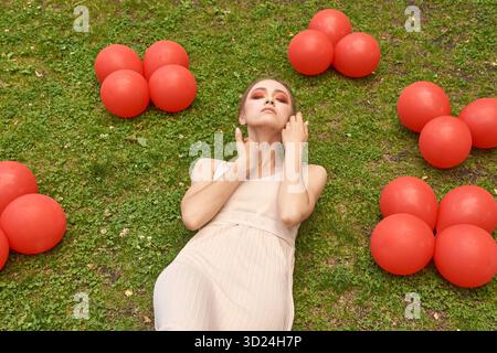 Giovane donna caucasica che si rilassa sull'erba circondata da palloncini rossi all'aperto Foto Stock