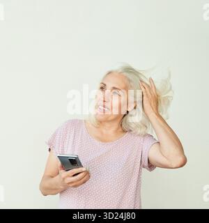 Anziana donna caucasica con il telefono che regola i capelli, sorridente, in luce informale Foto Stock