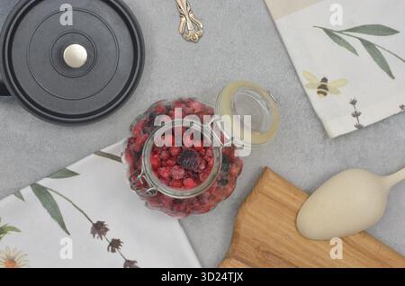 Ribes rosso e nero congelati in vaso decorativo, asciugamano da cucina, cucchiaio, coperchio del recipiente nero e tagliere. Tavolo da cucina vintage in marmo. Foto Stock
