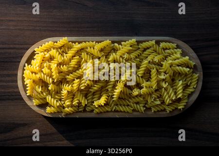 Pasta rotini cruda in una ciotola di legno su un tavolo di legno scuro sotto la luce naturale del sole. Foto Stock