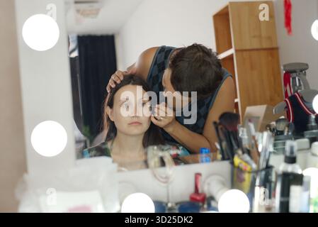 Giovane donna caucasica adulta seduta mentre il giovane adulto caucasico applica il trucco al viso davanti allo specchio, vari prodotti cosmetici e strumenti visibili sul tavolo Foto Stock