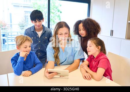Un fisioterapista pediatrico guida un gruppo di bambini durante una sessione di riabilitazione, utilizzando una compressa per riesaminare gli esercizi. Ambientalisti calorosi e solidali Foto Stock