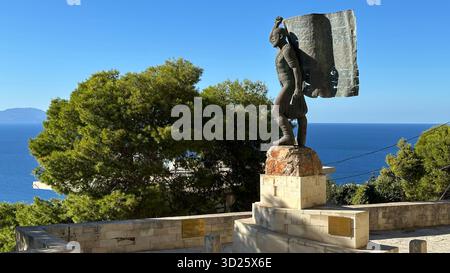 Eroica statua in bronzo di Spyros Kayales che innalza la bandiera greca al parco Venizelos Graves di Akrotiri, Chania, Creta, Grecia, con sfondo marino. Foto Stock